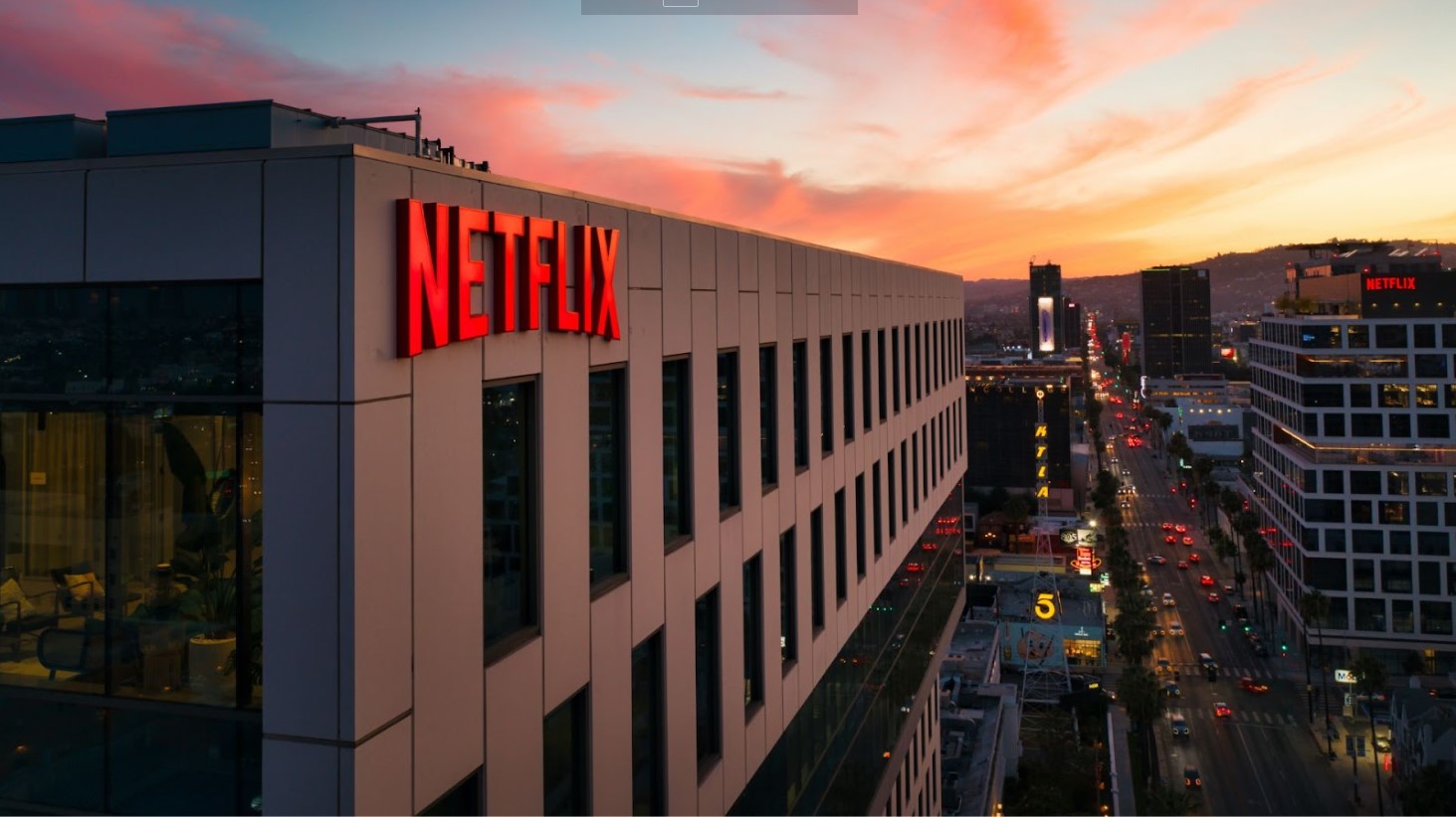 The Netflix logo illuminated in red on the side of a modern office building during sunset, with the cityscape of Los Angeles and streets with moving traffic in the background