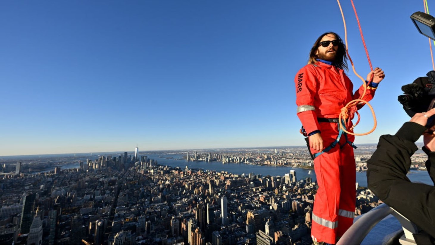 Jared Leto, wearing a bright orange jumpsuit with 'MARS' written on one sleeve, stands high above New York City atop the Empire State Building