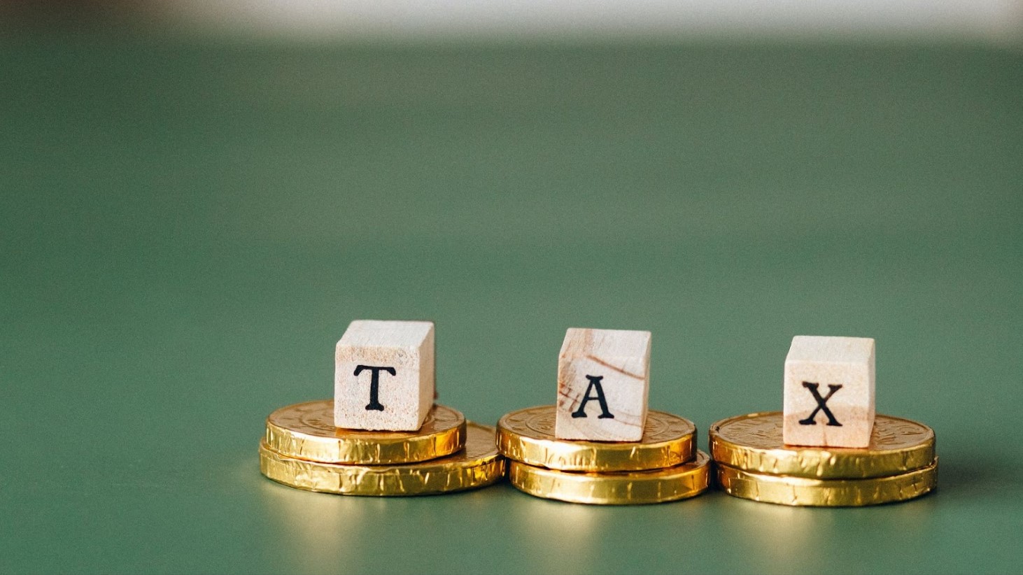 The word "TAX" is spelled out with individual letter blocks placed on top of stacked golden coins against a soft green background