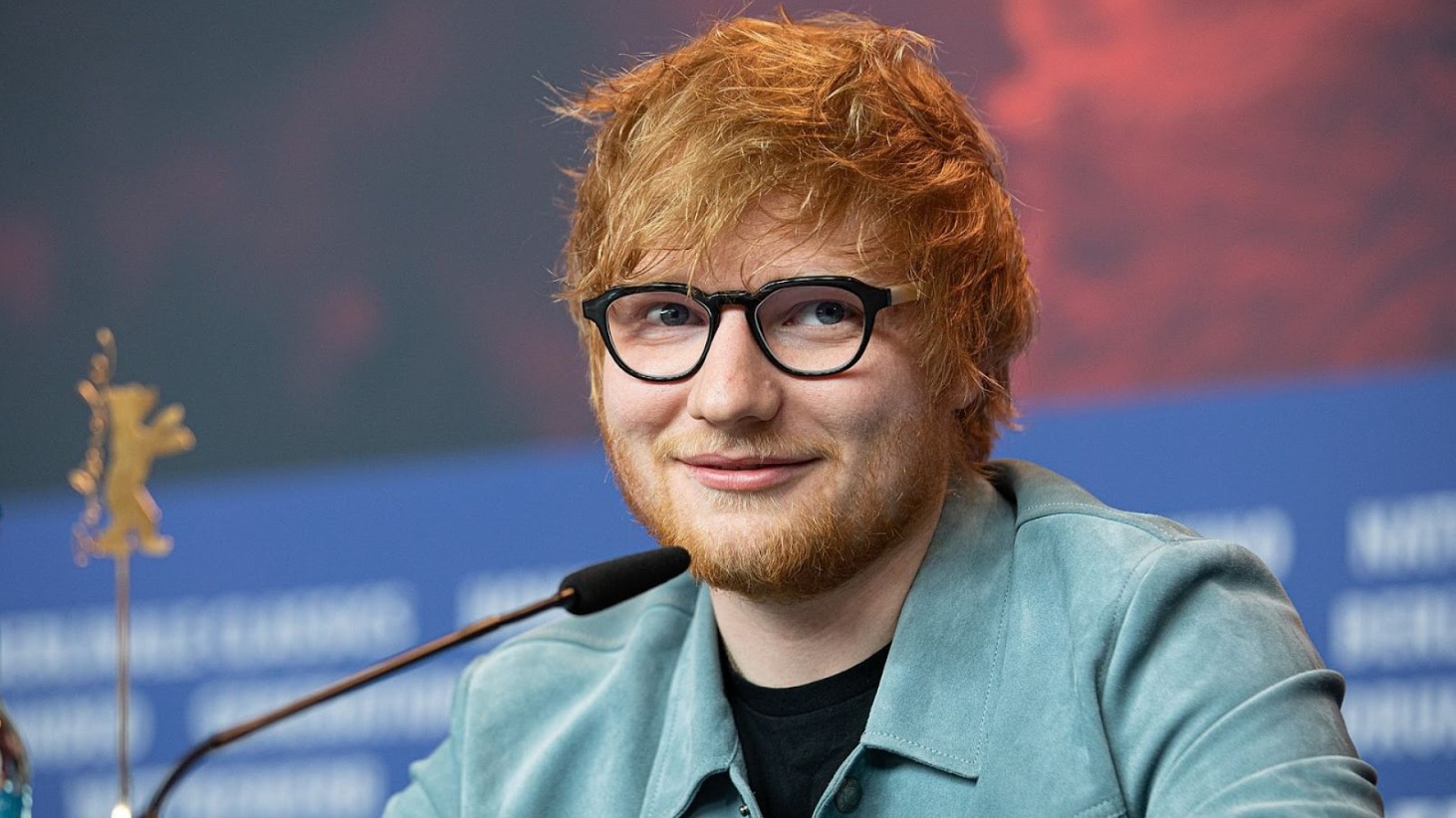 Ed Sheeran is smiling behind a microphone at a press conference. He's wearing black-rimmed glasses and a blue blazer over a black shirt