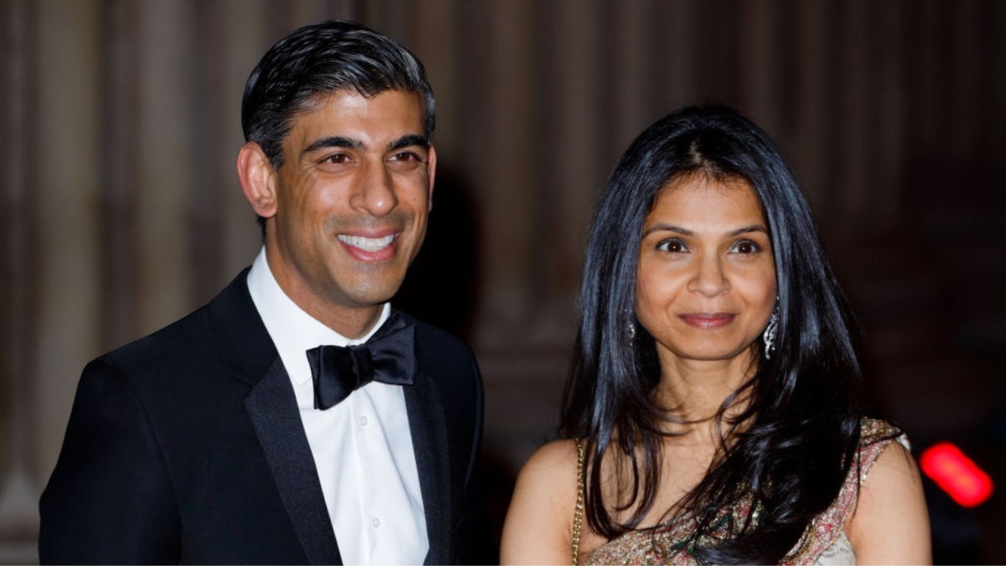 Rishi Sunak and Akshata Murty are photographed together at a formal event. The man is wearing a classic black tuxedo with a bow tie and has a cheerful smile. The woman beside him is wearing a traditional Indian dress with intricate embroidery and is also smiling