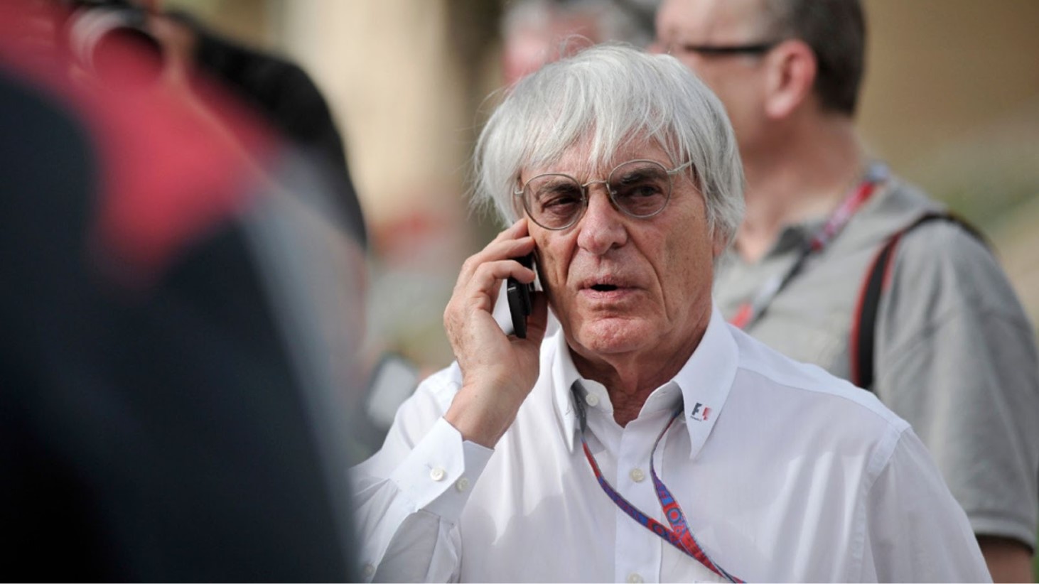 Bernie Ecclestone wearing a white shirt and a lanyard, is speaking on a mobile phone