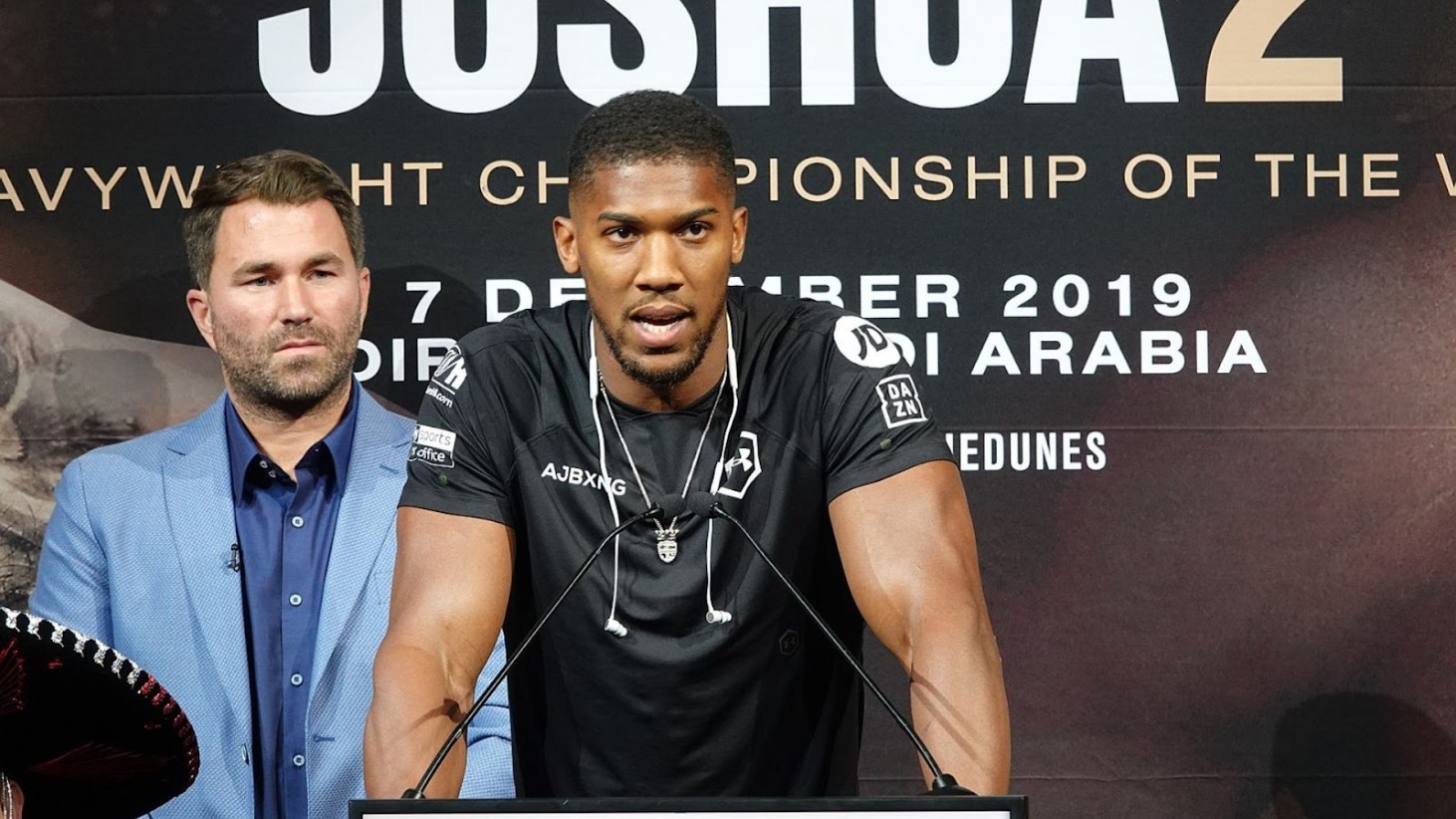 Anthony Joshua stands at a podium during a press conference. He is wearing a black T-shirt with the letters "AJBXNG" printed on it