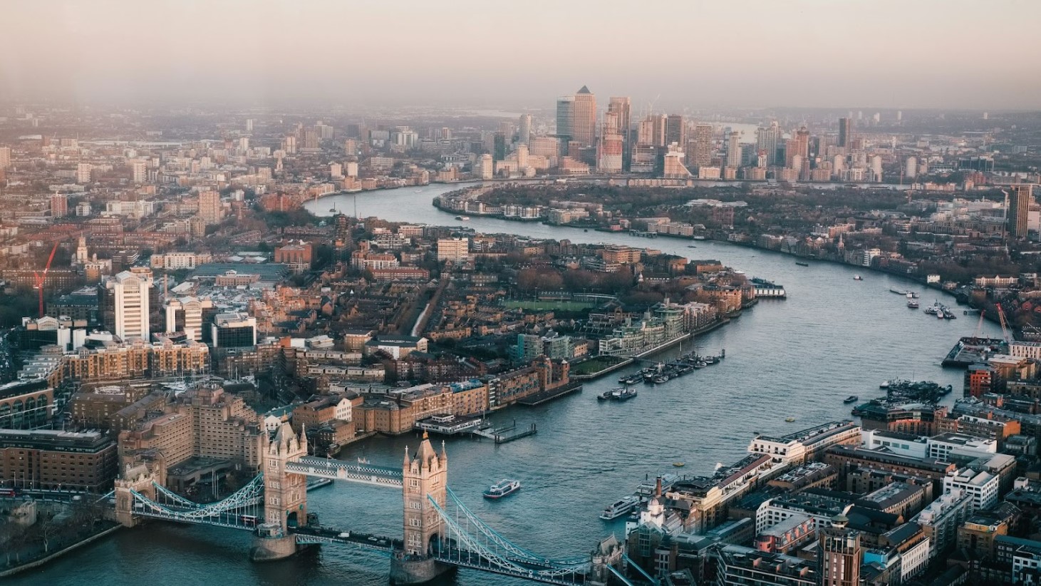 A photograph captures a sprawling aerial view of London, with the Thames River winding through the city. The Tower Bridge is prominent in the foreground, and the Canary Wharf district's skyscrapers can be seen in the distance