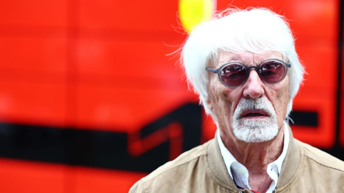 Bernie Ecclestone is pictured wearing sunglasses and a light brown jacket. He stands in front of a red background with a blurred black pattern
