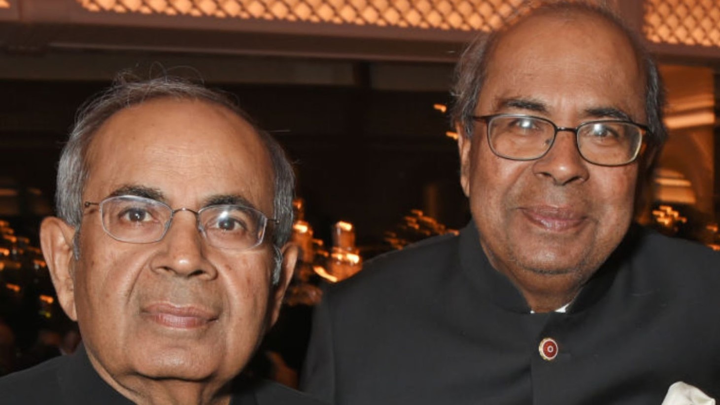 Two older men are photographed up close, smiling at a formal event. Both are wearing traditional Indian attire and glasses