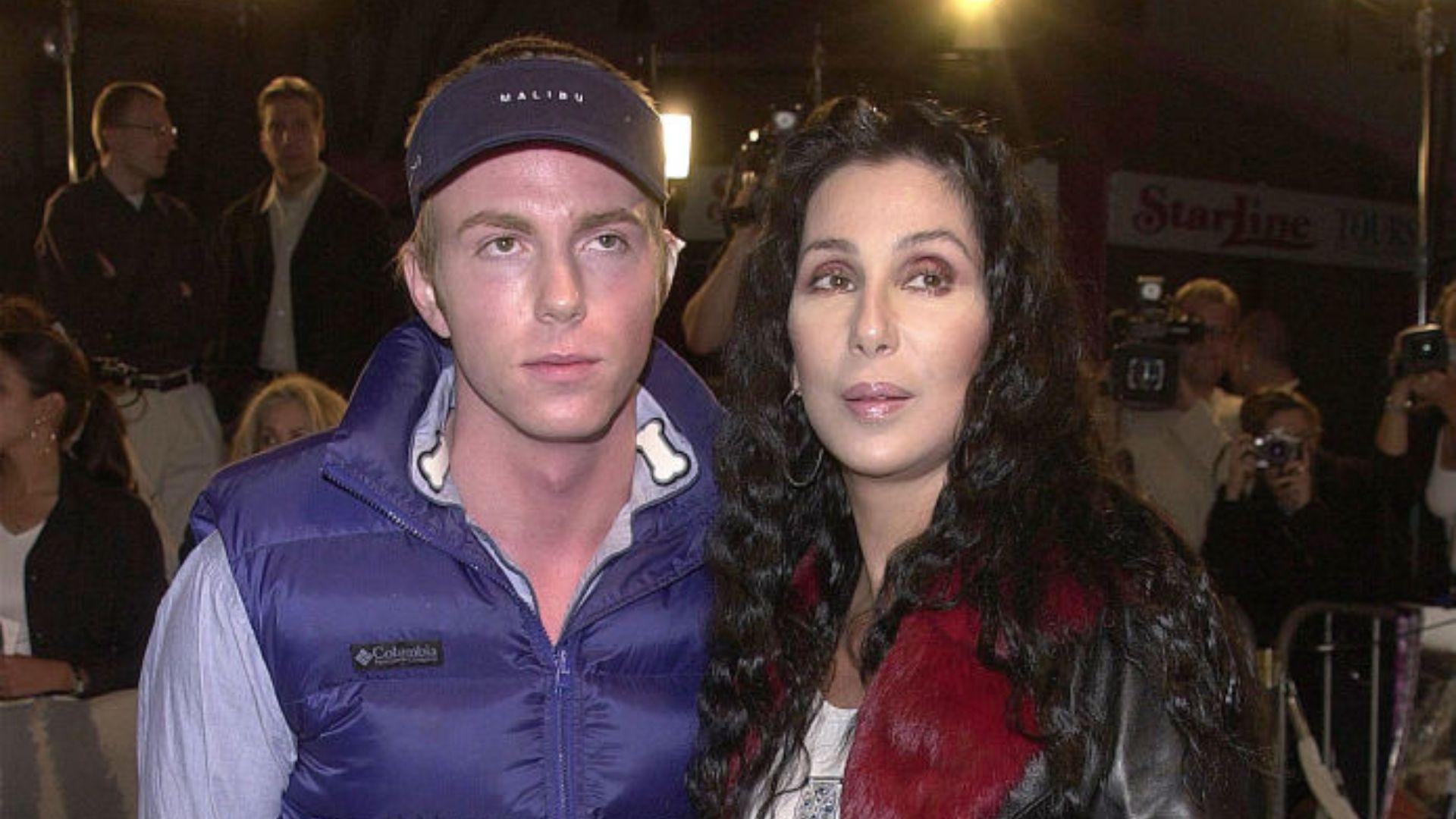 Cher and Elijah posing together at a public event. The man is wearing a blue and white Columbia jacket and a dark cap with the word "Malibu" on it, while the woman to his right is wearing a brown leather jacket with red fur trim