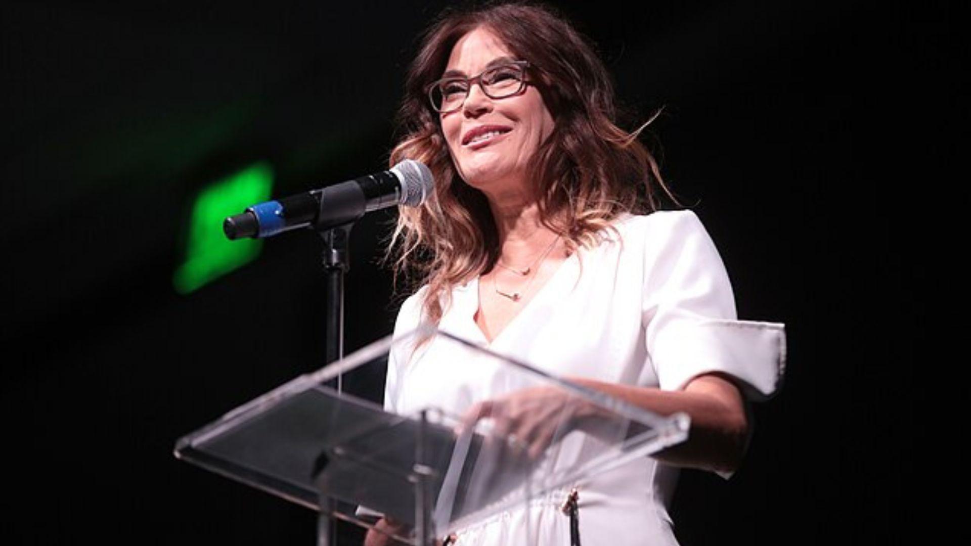 Teri Hatcher giving a talk. She is wearing a light pink dress and glasses and is standing behind a glass stand that has some paper and a microphone.