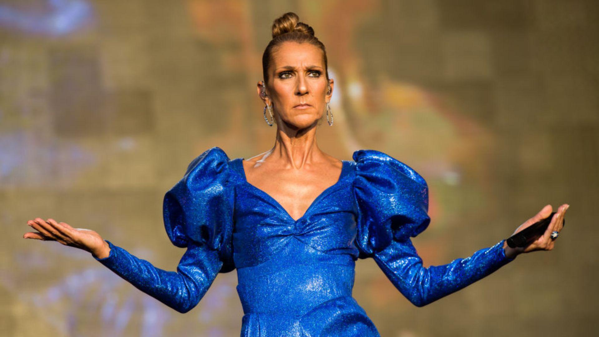 Céline Dion wearing a shimmering blue metallic dress with pronounced shoulder puffs and a plunging neckline. Her hair is styled in a tight bun, and she accessorizes with large hoop earrings